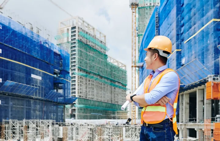 Certified construction safety professional standing with crossed arms in front of a construction site with scaffolding.